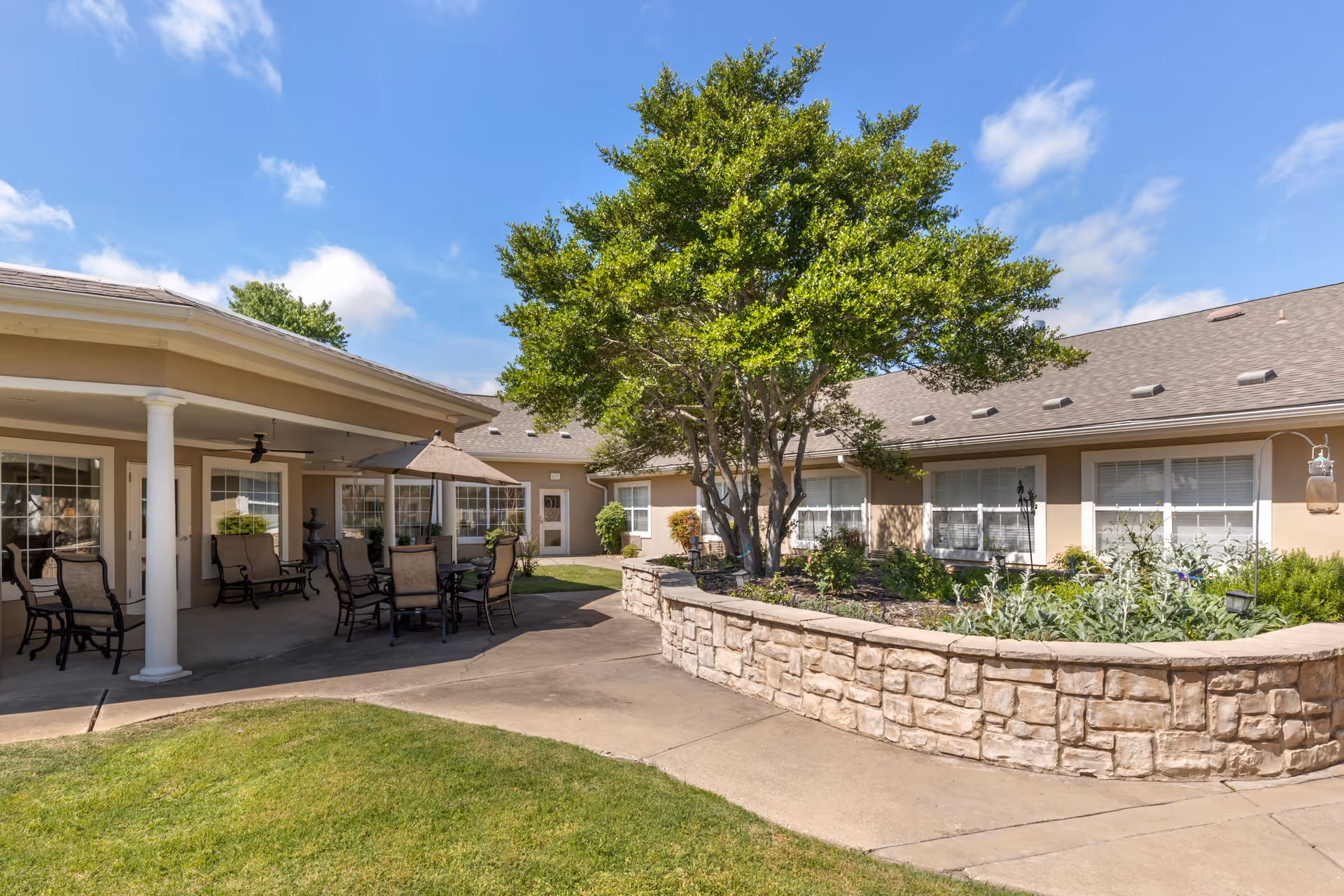 Outdoor patio area at Brookdale Creekside featuring a covered seating area with chairs and a table under an umbrella, a stone planter with a tree and various plants, and a building with multiple windows in the background under a blue sky with some clouds.