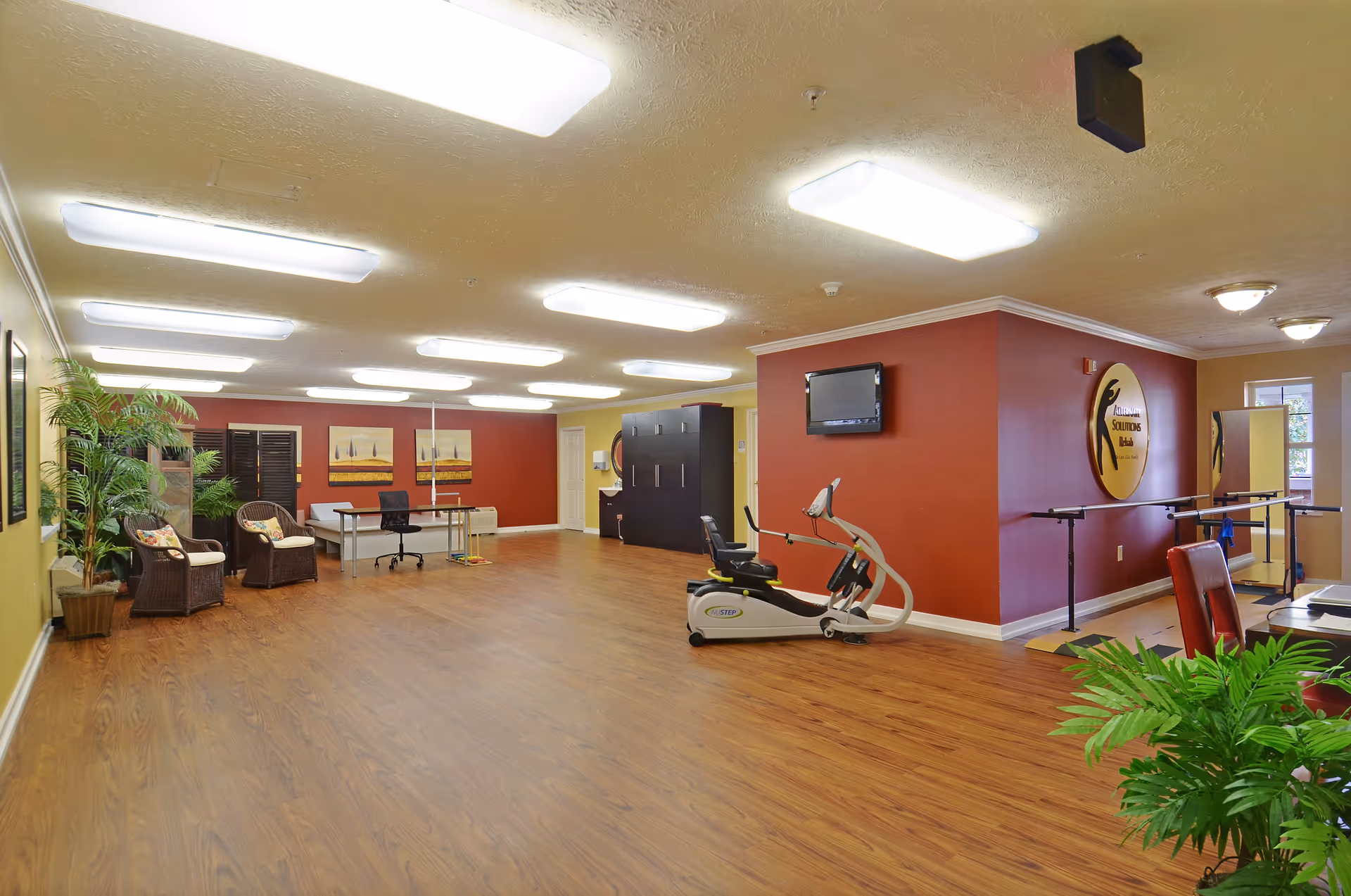 A spacious senior living facility room with wood flooring and bright overhead lighting. The room features exercise equipment, wicker chairs with cushions, a desk with a chair, potted plants, and a wall-mounted TV. The walls are painted in warm tones of red and yellow, and there is a large circular sign on one wall that reads 'Stoney Ridge Senior Living'.