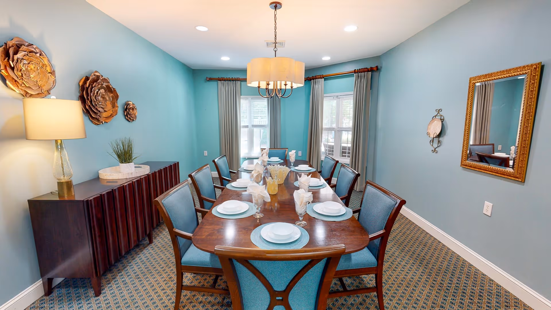 A dining room with a long wooden table set for eight people with white plates, glasses, and folded napkins. The room has blue walls, a patterned carpet, a sideboard with a lamp and decorative items, and a large window with curtains. A chandelier hangs above the table, and a gold-framed mirror is on one wall.