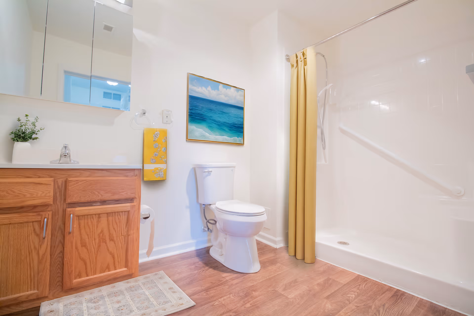 A clean and bright bathroom with wooden cabinets under a sink, a toilet, a shower with a yellow curtain, a wall-mounted grab bar, a small plant on the counter, a yellow towel hanging on the wall, and a framed picture of an ocean scene above the toilet.
