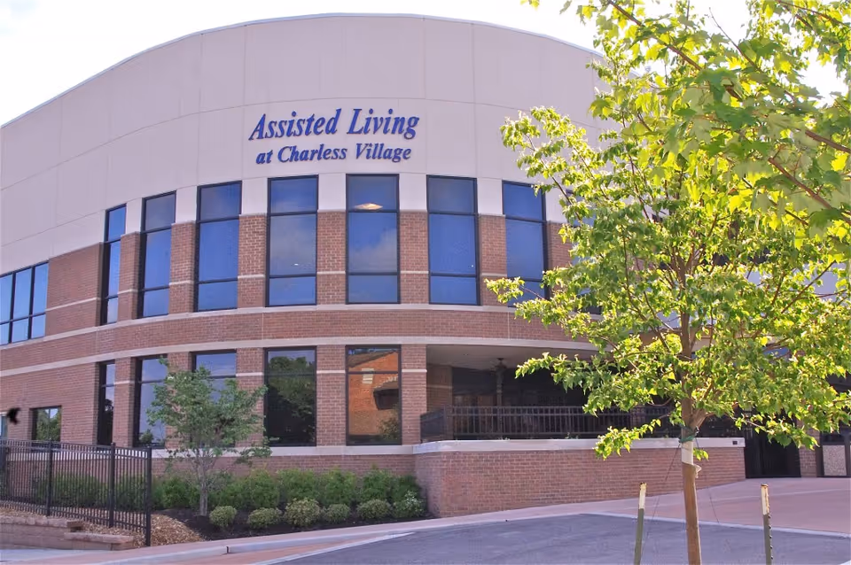 Exterior view of a two-story assisted living facility building with large windows, brick and beige walls, a small landscaped area with bushes and trees, and a sign that reads 'Assisted Living at Charless Village'.