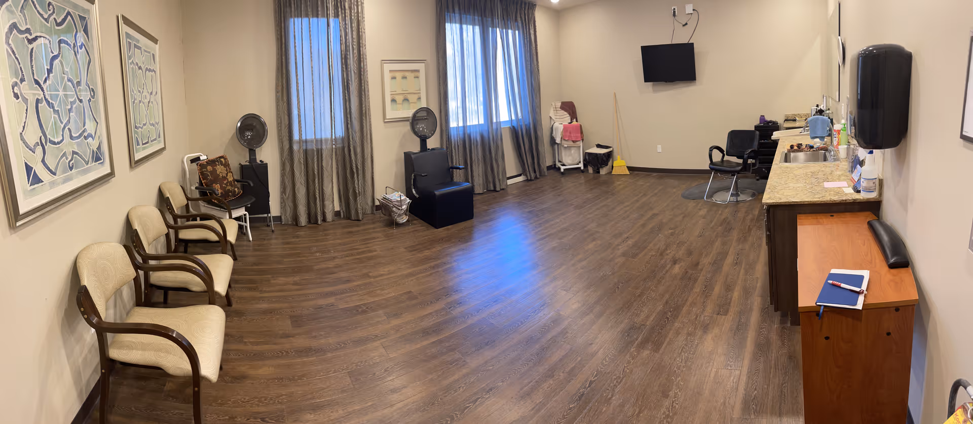 A spacious room with wood flooring and beige walls featuring several chairs lined up along the left wall, two windows with curtains, a wall-mounted TV, a countertop with a sink and various items, and a small wooden cabinet with a notebook and pen on top. The room appears clean and organized with some cleaning supplies visible in the corner.