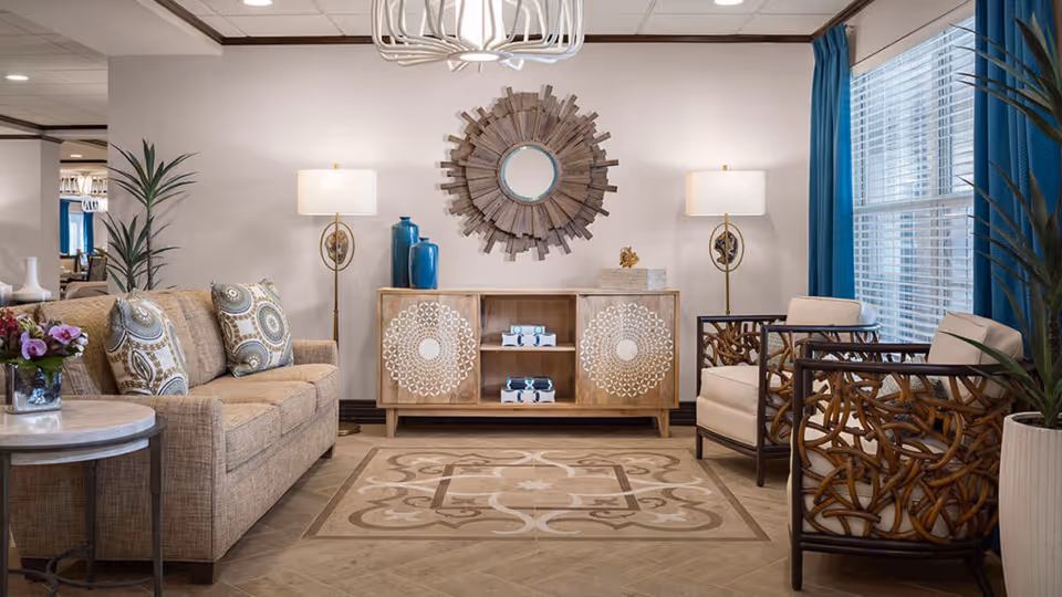 A cozy and stylish living room with a beige sofa adorned with patterned cushions on the left, two decorative wooden armchairs with cream cushions on the right, a wooden cabinet with intricate white designs in the center, topped with blue vases and decorative items. Two floor lamps flank the cabinet, and a round wooden sunburst mirror hangs above it. The room features a patterned area rug, a small round side table with a flower arrangement, large windows with blue curtains, and potted plants adding greenery.
