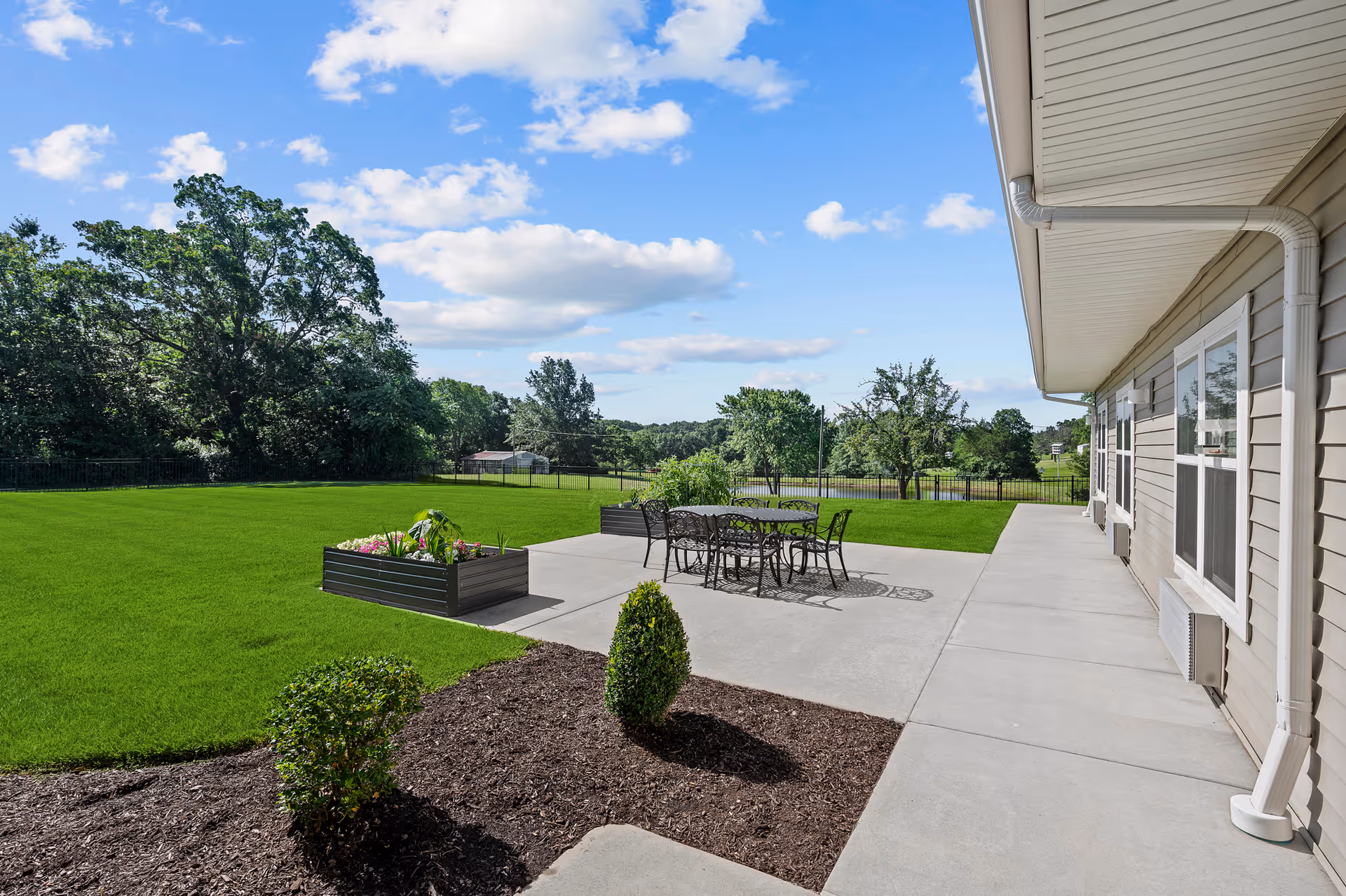 Concrete patio and walkway beside a senior living building overlooking a grassy yard with patio furniture, planter boxes, and trees.