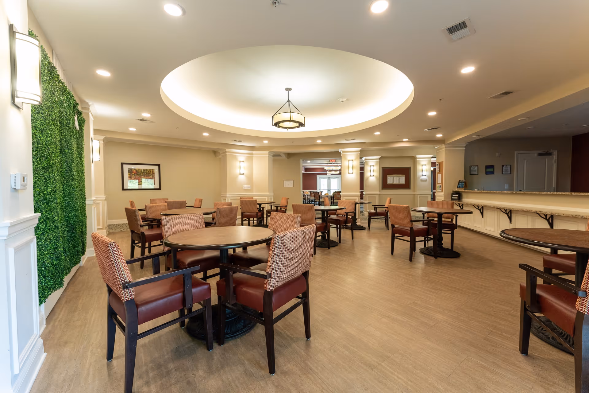 A spacious dining room with multiple round tables and cushioned chairs arranged neatly. The room features a large circular recessed ceiling light fixture, wall sconces, and a green vertical garden on one wall. The floor is wooden, and there is a long counter on the right side of the room.
