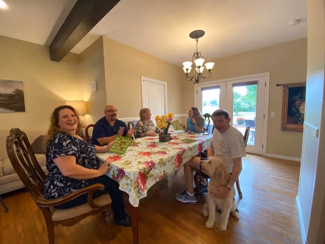Five people sitting around a dining table covered with a floral tablecloth in a well-lit room. One person is petting a large dog sitting on the floor. The room has wooden floors, a chandelier, and glass doors leading outside.