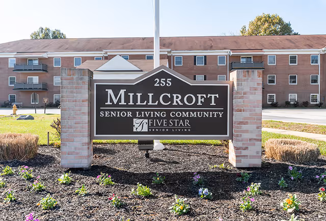 Outdoor view of the Millcroft Senior Living Community sign with a brick apartment building in the background, surrounded by a landscaped area with flowers and mulch.