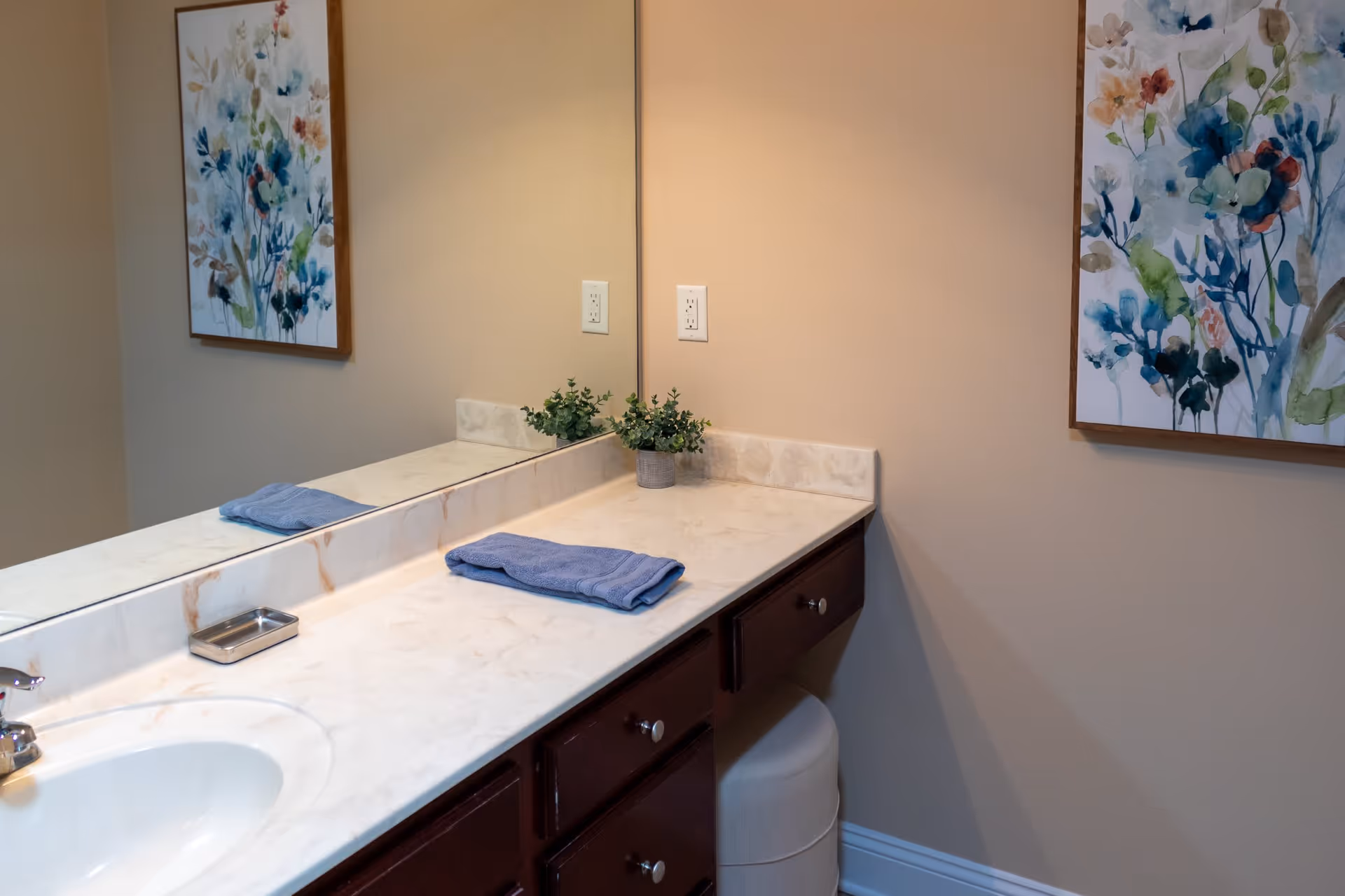 Bathroom countertop with a sink, a folded blue towel, a small potted plant, and a soap dish. A large mirror runs along the wall above the countertop, reflecting a floral painting on the opposite wall. The cabinetry below the countertop is dark wood, and the walls are painted beige.