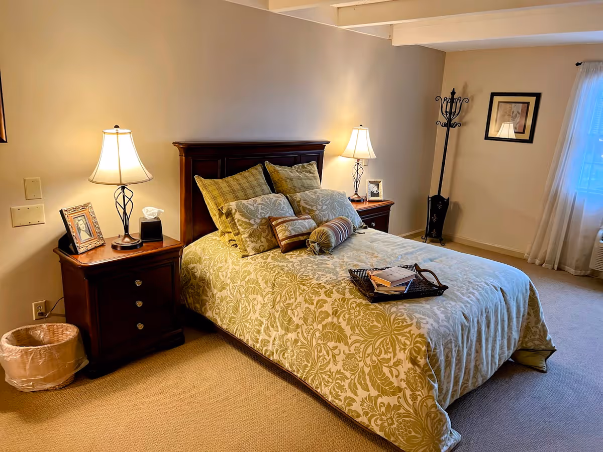A cozy bedroom with a double bed featuring a patterned bedspread and multiple pillows. There are two wooden nightstands on either side of the bed, each with a lamp and a framed photo. A coat rack stands in the corner near a window with sheer curtains, and a small trash bin is placed beside one nightstand.