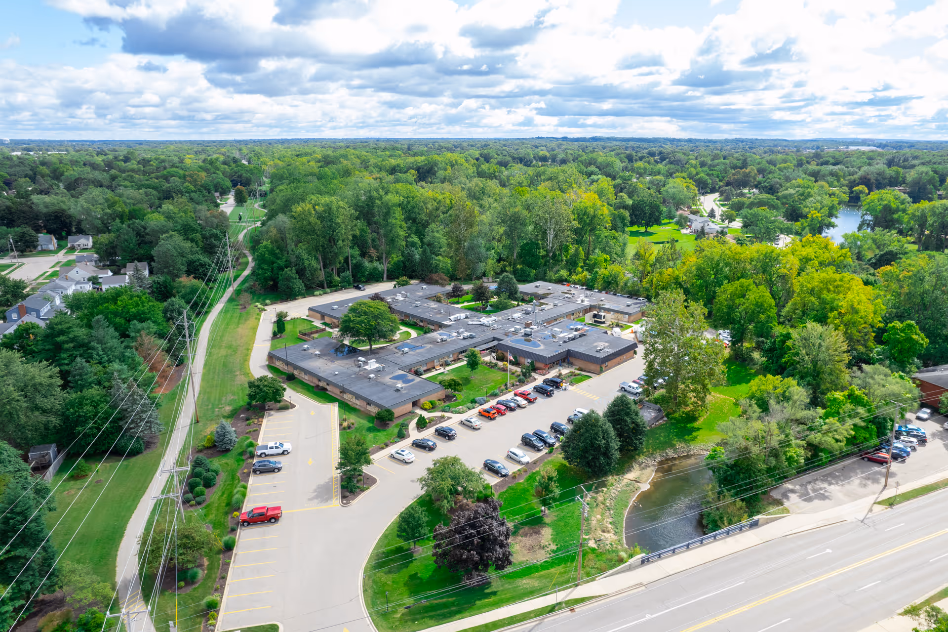 Aerial view of a single-story care facility complex with a parking lot, surrounding trees, and a small pond beside a road.