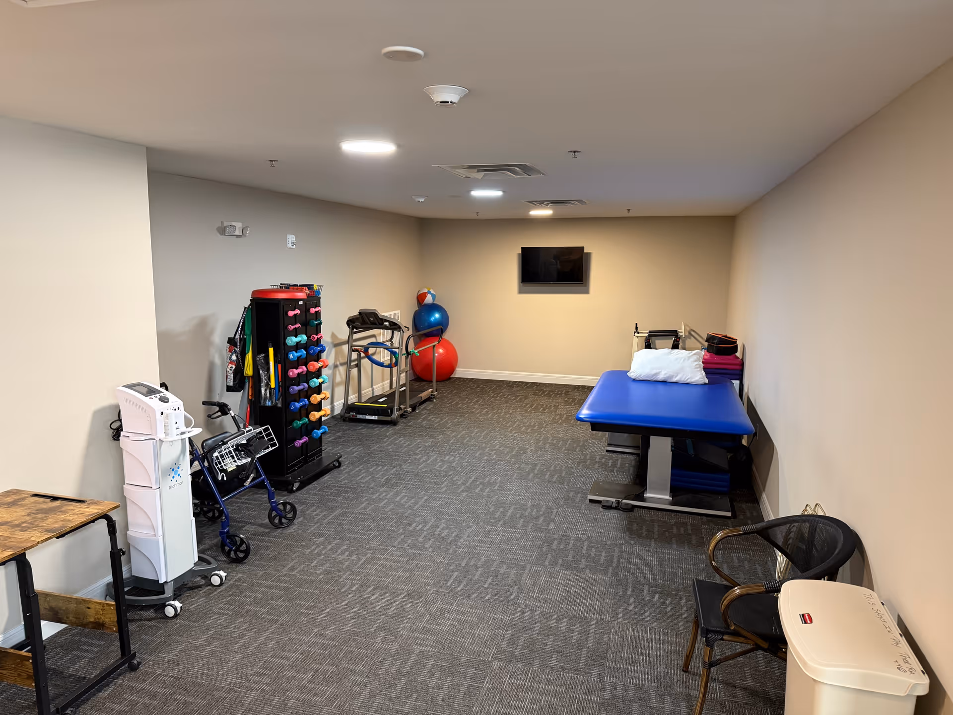 A senior living facility exercise room with a blue therapy table, a rack of colorful dumbbells, exercise balls, a treadmill, a walker, a chair, and a small table. The room has beige walls, gray carpet, and a wall-mounted TV.