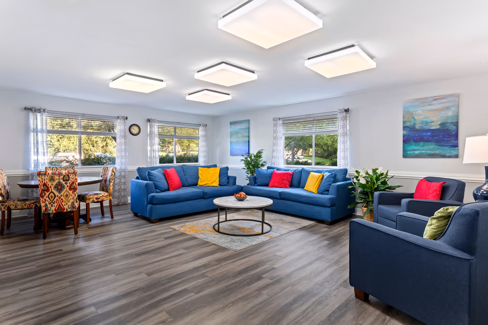 Bright and spacious living room with two blue sofas and two blue armchairs, each adorned with colorful throw pillows in red, yellow, and green. A round coffee table sits on a patterned rug in the center. Large windows with sheer curtains let in natural light, and two abstract paintings hang on the walls. A small round dining table with four patterned chairs is visible in the corner. The room has wood flooring and modern square ceiling lights.
