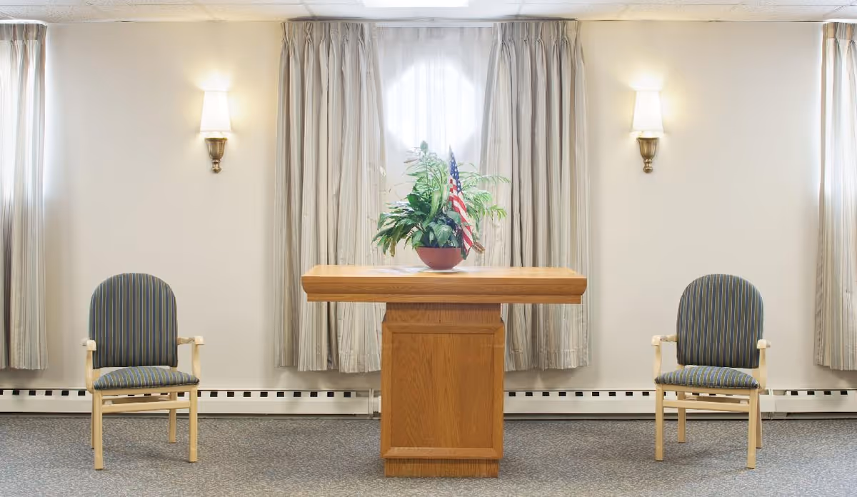 A simple room with two striped armchairs placed on either side of a wooden podium table. Behind the podium is a window with light-colored curtains, and a potted plant with an American flag is on top of the podium. The walls are light-colored with two wall sconces providing soft lighting.