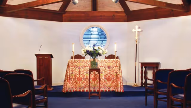 Interior view of a small chapel with wooden ceiling beams, a round stained glass window, an altar covered with a decorative cloth, a vase of flowers on a stand, a lectern, a tall candle stand with a cross, and several wooden chairs arranged facing the altar.