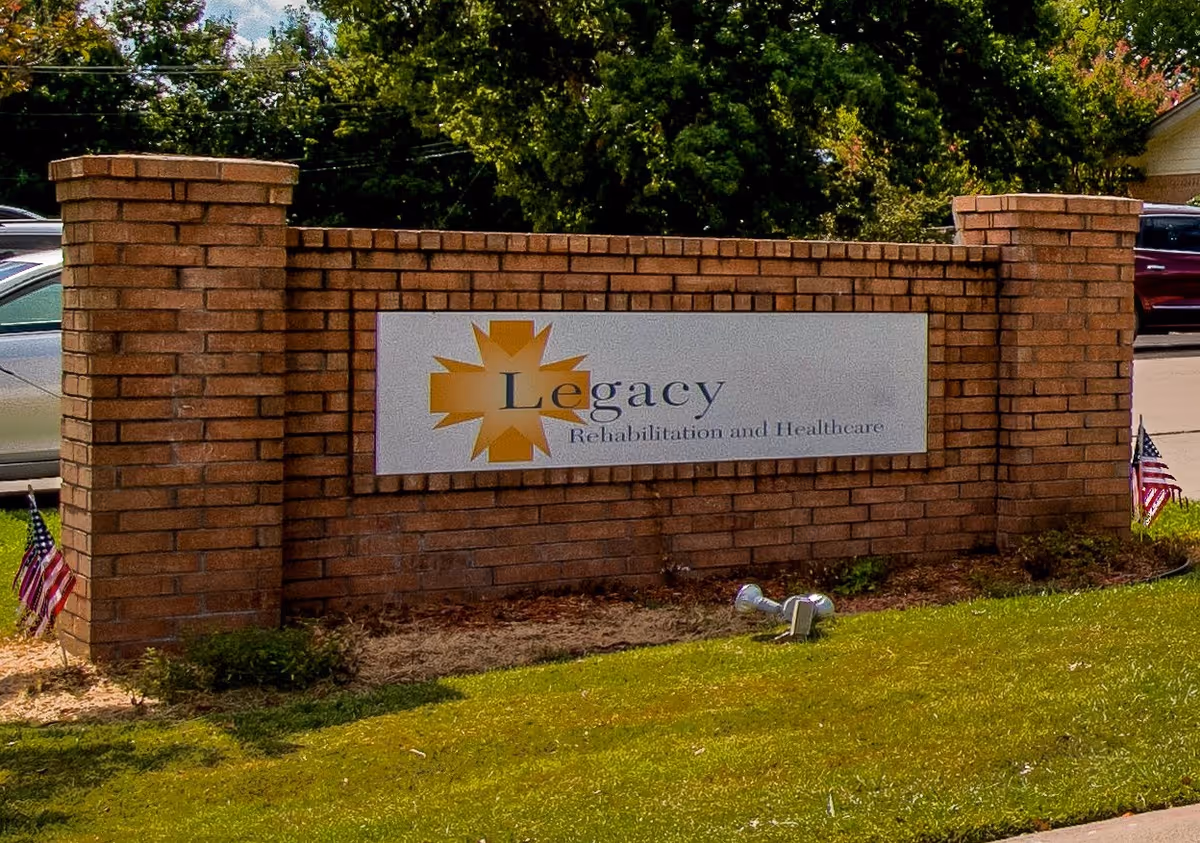 A brick sign for Legacy Rehabilitation and Healthcare mounted on a low brick wall with two American flags on either side, surrounded by grass and some bushes, with trees and parked cars in the background.