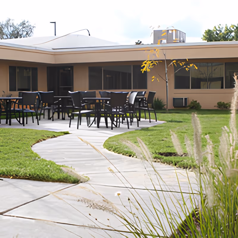 Curved concrete path leading to an outdoor patio area with tables and chairs in a grassy courtyard next to a single-story building.