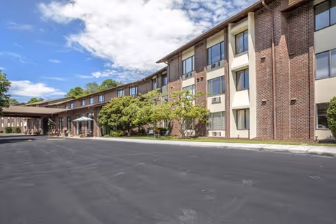 Front exterior of a curved brick senior living building with a covered entrance, three-story windows, landscaping, and a paved driveway.