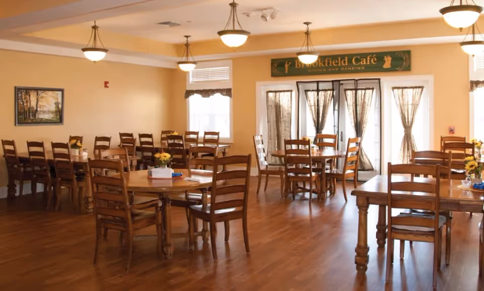 A dining room with wooden tables and chairs arranged neatly on a wooden floor. The room has soft yellow walls, ceiling lights, and windows with sheer curtains. A sign above a set of glass doors reads 'Brookfield Café Dining and Dancing'. Small flower arrangements are placed on the tables.
