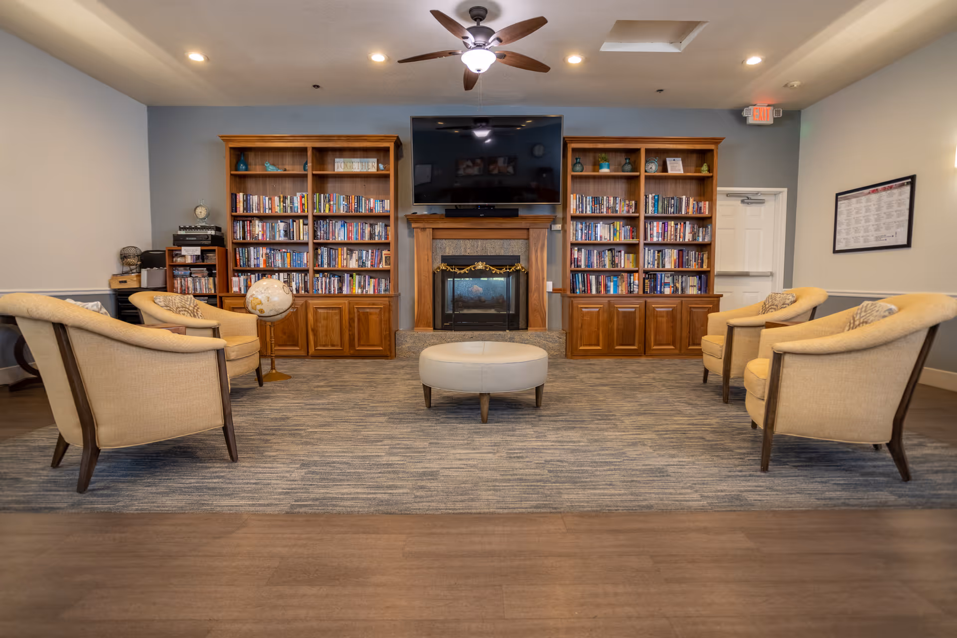 A comfortable senior living common room with bookshelves, a fireplace and TV, and chairs arranged around a central ottoman.