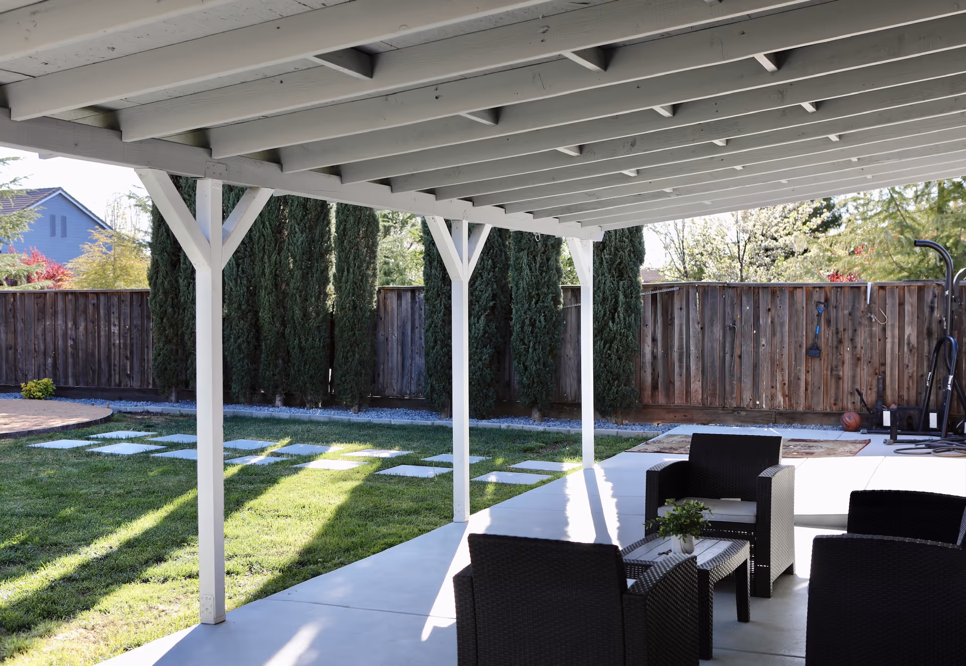 Covered patio with wicker seating and table overlooking a grassy backyard with stepping stones and a wooden fence.
