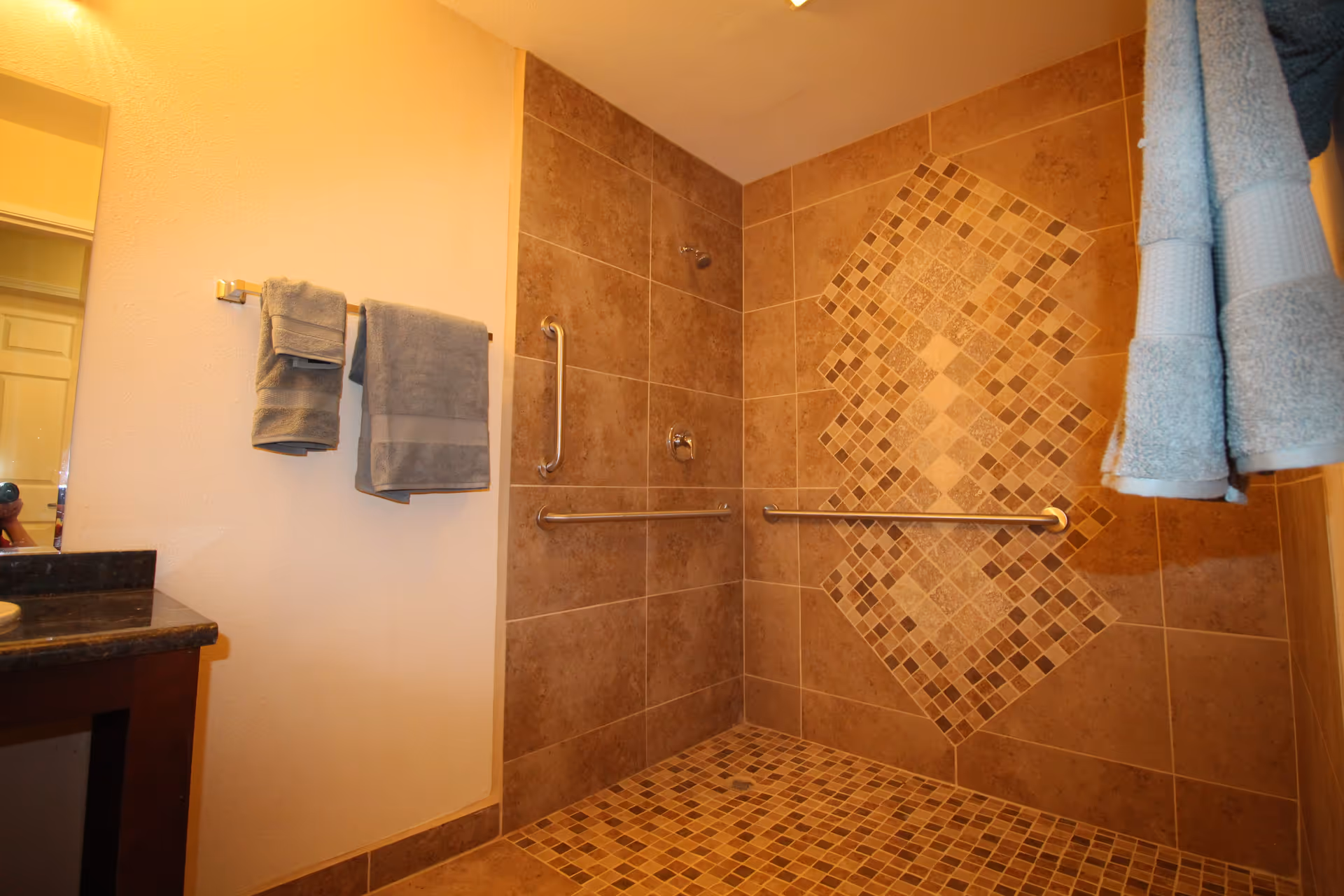 A bathroom shower area with brown and beige tiled walls and floor. The shower has a decorative diamond pattern of smaller tiles on the wall. There are two metal grab bars installed on the walls for support. Towels hang on a rack to the left and on hooks to the right. A portion of a dark countertop and mirror is visible on the left side of the image.