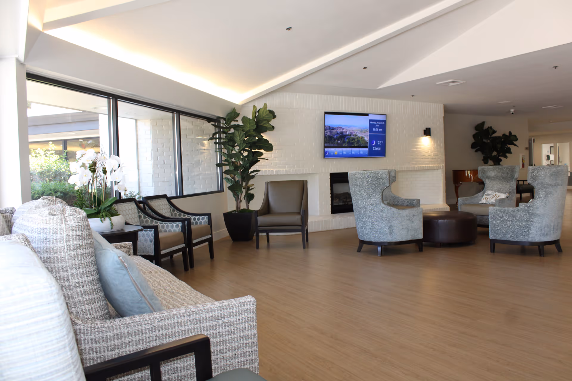 A spacious and well-lit living room area in a senior living facility with a variety of seating options including cushioned armchairs, a sofa, and high-back chairs arranged around a central ottoman. The room features large windows letting in natural light, a white brick fireplace with a mounted flat-screen TV displaying weather information, and decorative plants placed around the space.