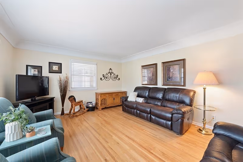 A cozy living room with a dark brown leather sofa, two green armchairs, a wooden cabinet, a flat-screen TV on a black TV stand, a floor lamp with a beige shade, and decorative wall art. The room has light-colored walls and wooden flooring.
