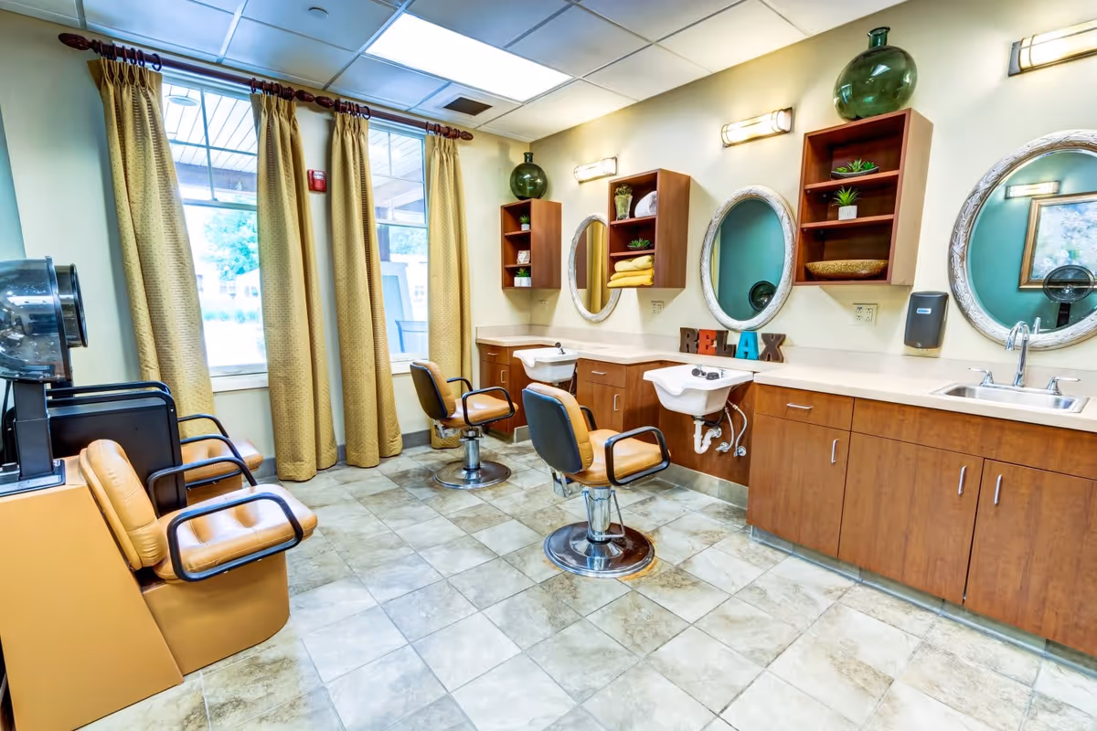 Interior of a salon area with three salon chairs in front of sinks and mirrors. There are wooden cabinets and shelves with decorative items, and large windows with yellow curtains letting in natural light. The word RELAX is displayed on the countertop.