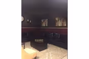 A dimly lit living room with dark walls and a maroon accent stripe. The room features black leather seating arranged around a central black ottoman, a beige armchair, a patterned area rug, and two abstract paintings on the wall.
