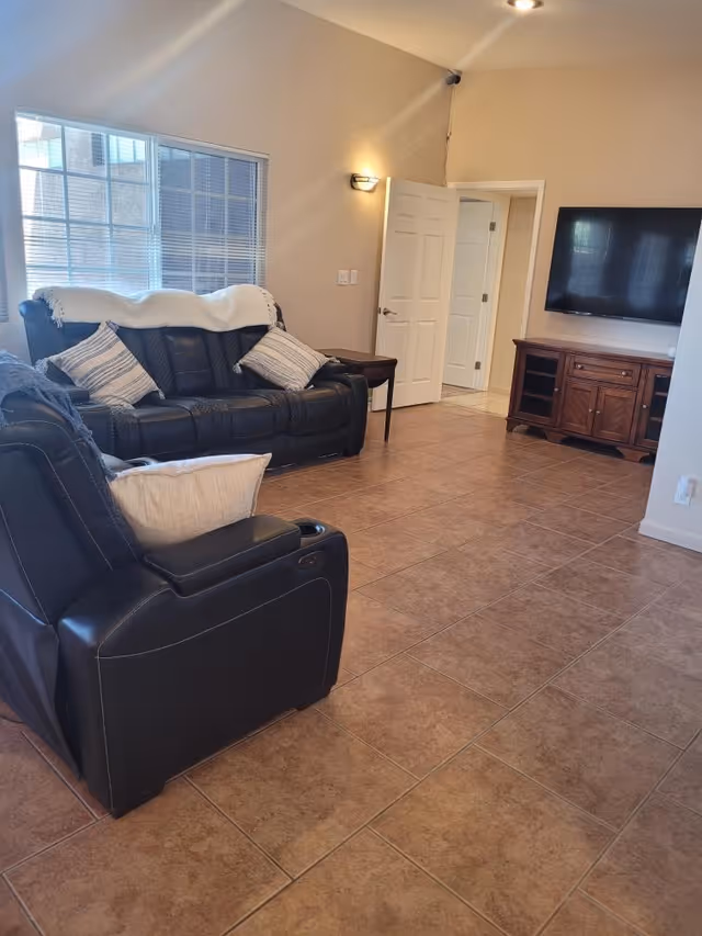 Bright living room with black leather seating, a wall-mounted TV on a wooden console, and tiled floors.