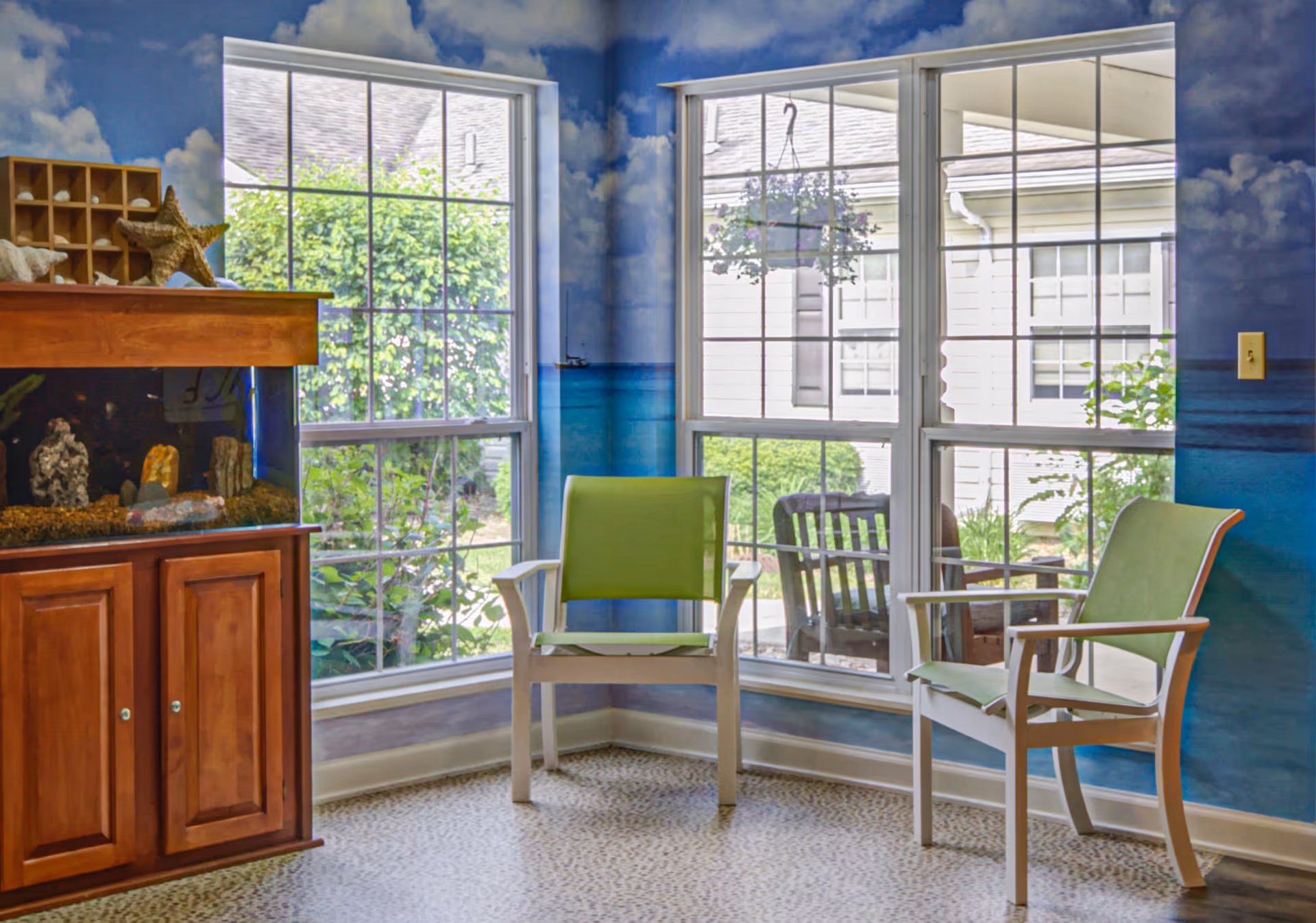 A corner of a room with two green chairs facing each other near large windows. The walls are painted with a mural depicting a blue sky with clouds and an ocean horizon. There is a wooden cabinet with an aquarium on top, decorated with seashells and a starfish. Outside the windows, greenery and parts of another building are visible.