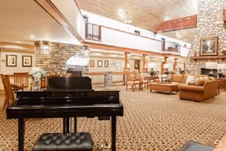 Spacious senior living common area with a black piano in the foreground, seating groups, a stone fireplace, and an upper mezzanine balcony.