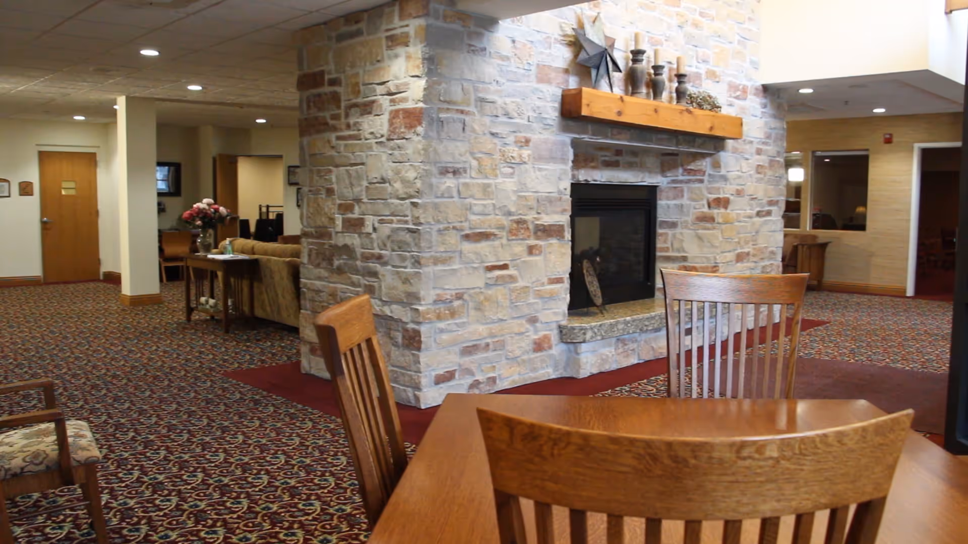 Interior view of a senior living facility lounge area featuring a stone fireplace with decorative items on the wooden mantel. In the foreground, there is a wooden table with chairs. The room has patterned carpet flooring and recessed ceiling lights. There is a seating area with a sofa and a table with a vase of flowers in the background.