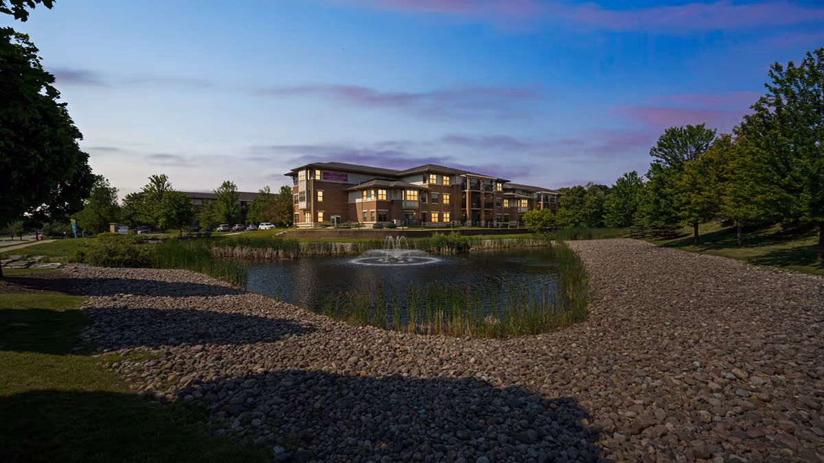 Exterior view of a senior living facility named Village at the Falls during dusk, showing a multi-story building with lights on, surrounded by trees and a landscaped area with a pond and a water fountain in the foreground.