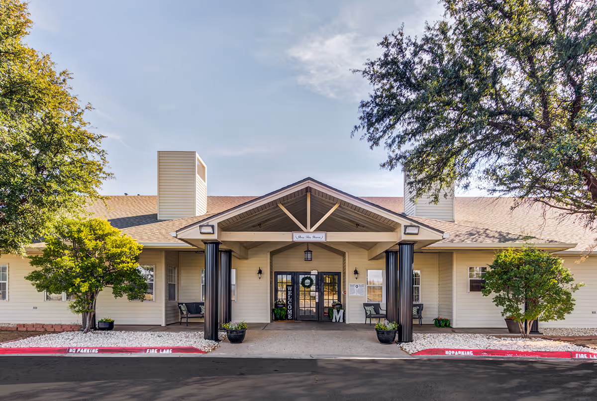 Front entrance of Marcy Place senior living building with a covered portico, columns, seating, and landscaped trees.