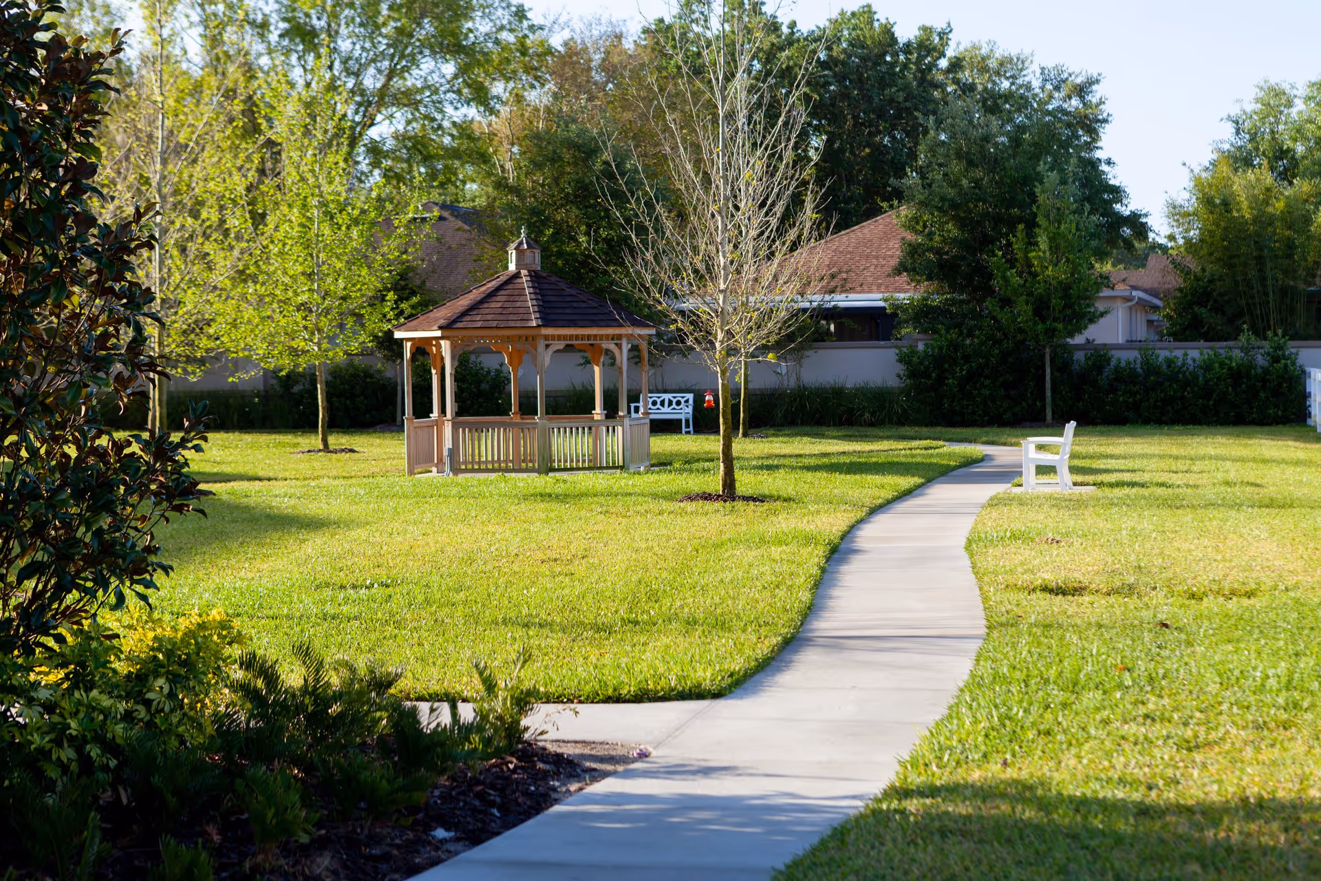 A serene garden scene featuring a wooden gazebo surrounded by lush green grass and trees, with a winding concrete path leading through the landscape.