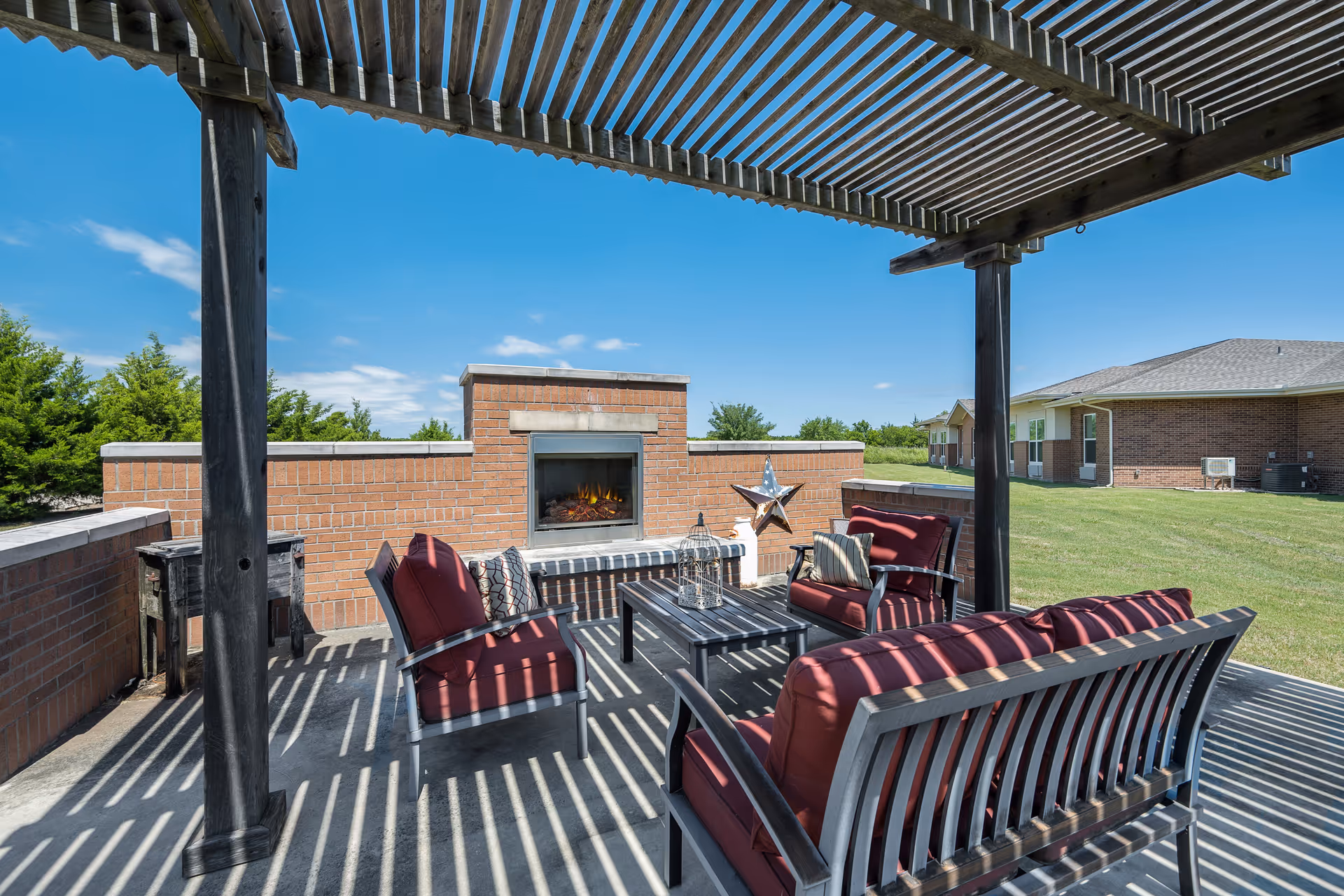 Outdoor seating area with cushioned chairs and a sofa under a wooden pergola, featuring a brick fireplace and a decorative star on the wall, with a grassy lawn and buildings in the background under a clear blue sky.