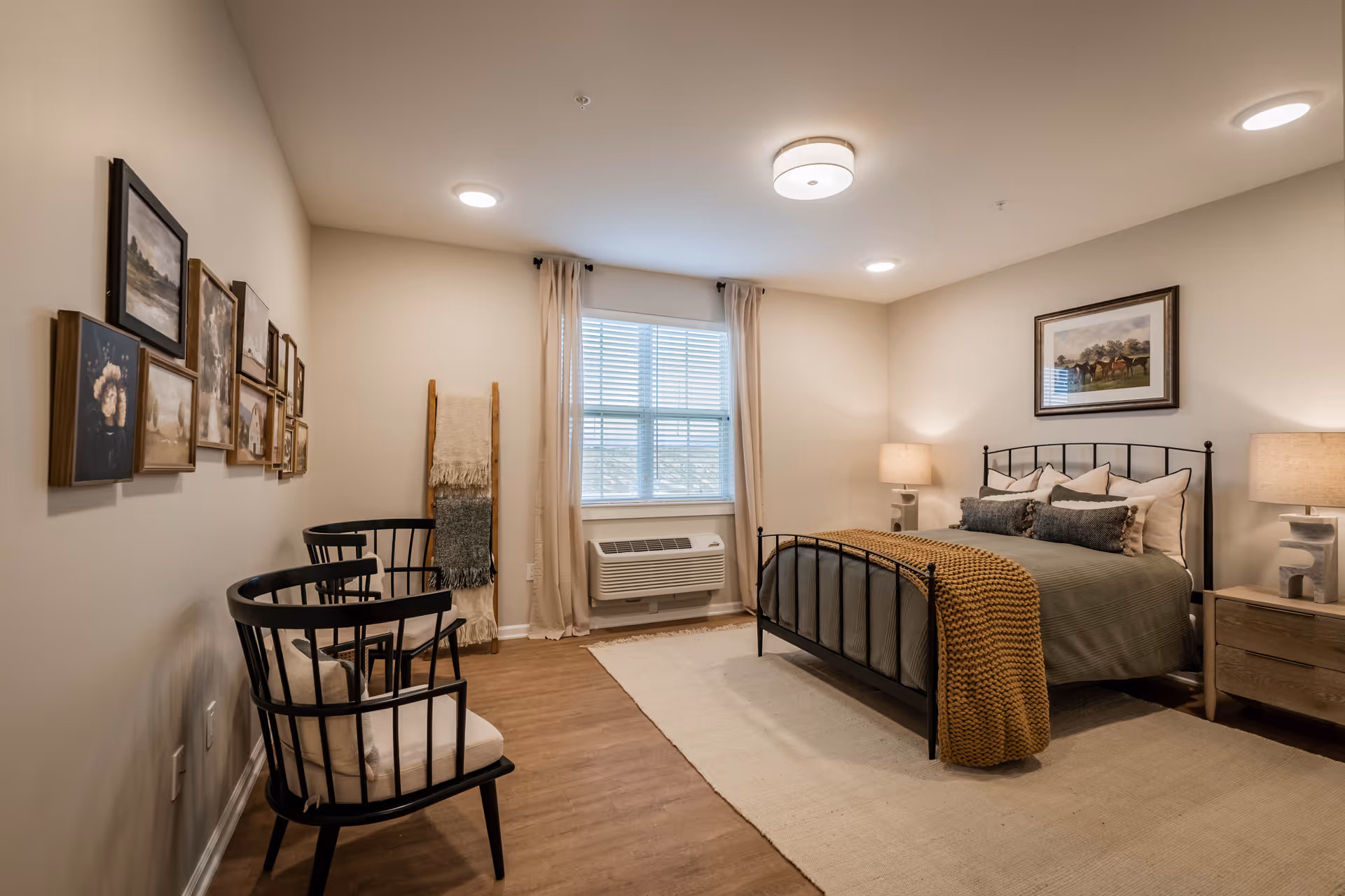 A cozy bedroom in a senior living facility featuring a bed with a black metal frame, green bedding, and a mustard-colored knitted throw. The room has two wooden nightstands with matching lamps, a window with beige curtains and blinds, two black cushioned chairs, a beige rug, and a wall decorated with framed artwork and a wooden ladder holding blankets.