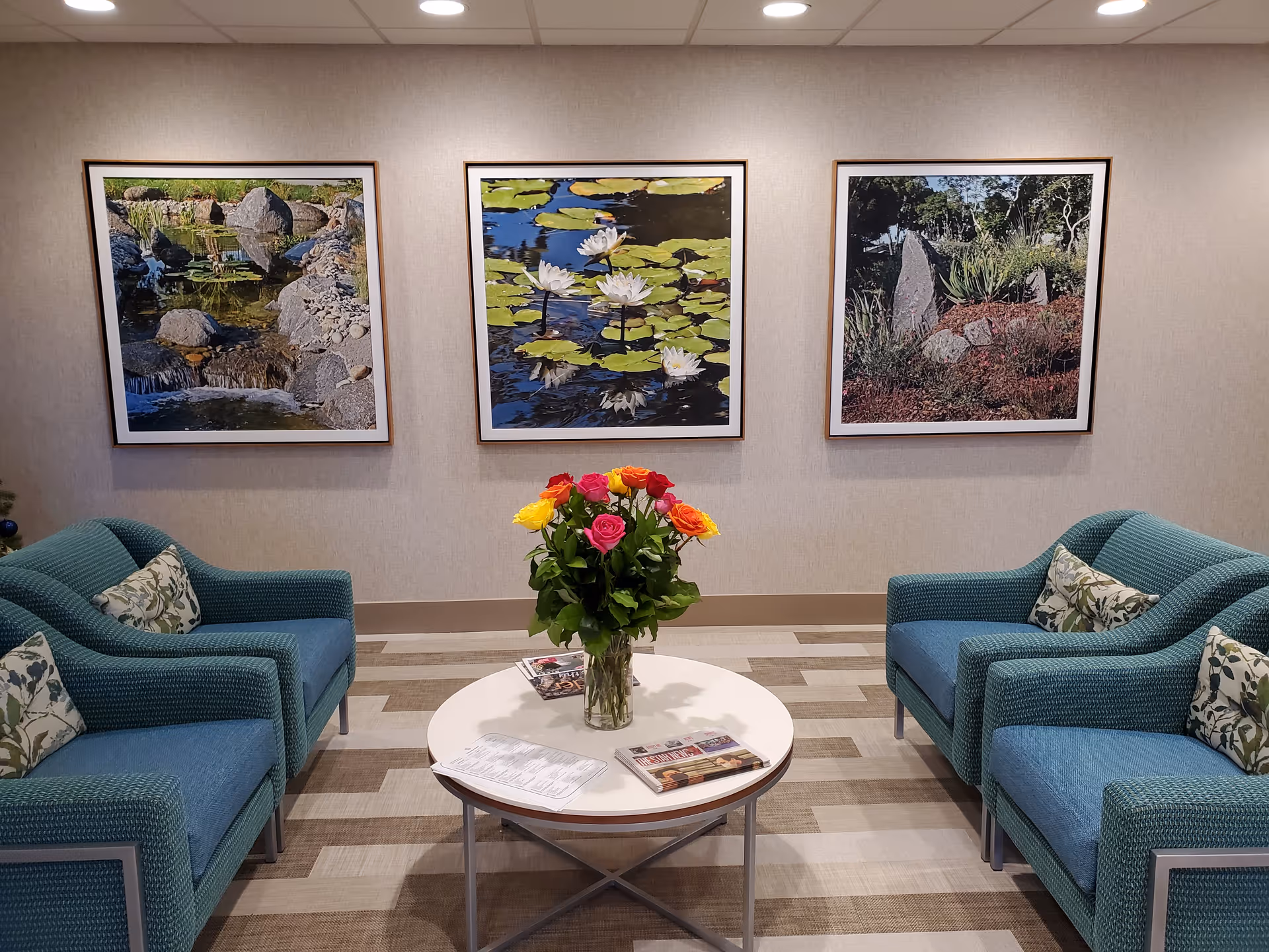 A cozy seating area with two teal armchairs facing each other, each with patterned cushions. Between them is a round white table with a vase of colorful roses and some magazines. On the wall behind the seating area are three framed nature photographs featuring rocks, water, and lily pads.