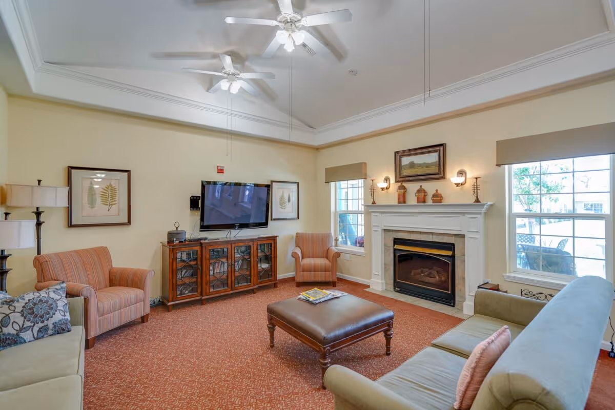 A cozy senior living community lounge with beige walls, two ceiling fans, a flat-screen TV mounted on the wall above a wooden cabinet, two striped armchairs, a green sofa with a floral pillow, a large brown leather ottoman, a fireplace with decorative items on the mantel, and two windows with beige valances letting in natural light.