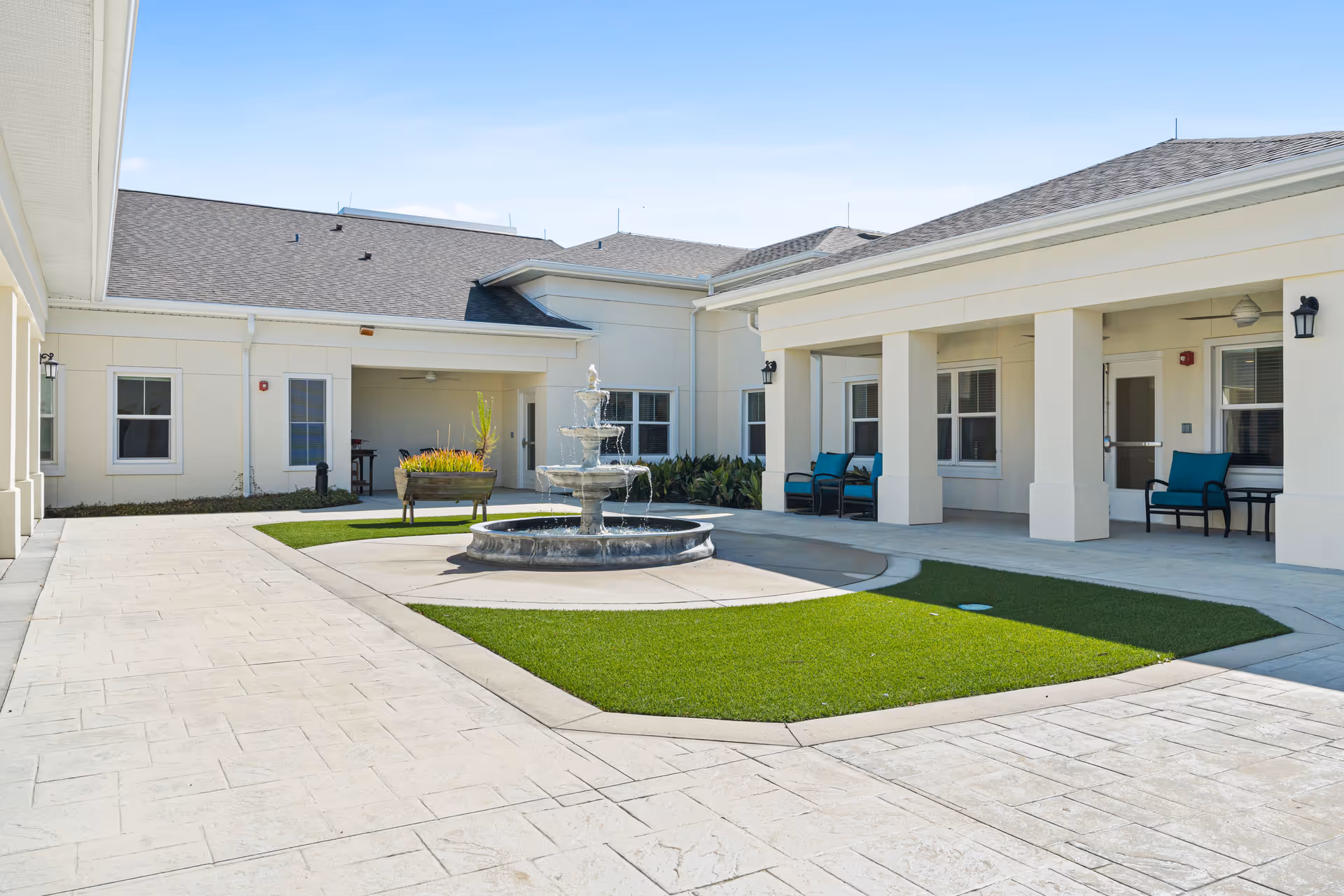 Outdoor courtyard area of a senior living facility with a central three-tiered water fountain surrounded by green artificial grass patches and beige tiled walkways. The building has white walls, multiple windows, and covered seating areas with blue cushioned chairs. The sky is clear and blue.