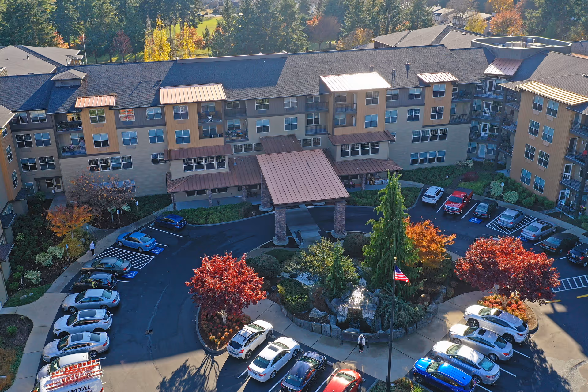 Aerial view of a senior living facility named Bonaventure of Lacey, showing a large multi-story building with a covered entrance, surrounded by a parking lot with several cars and landscaped areas with trees displaying autumn colors.