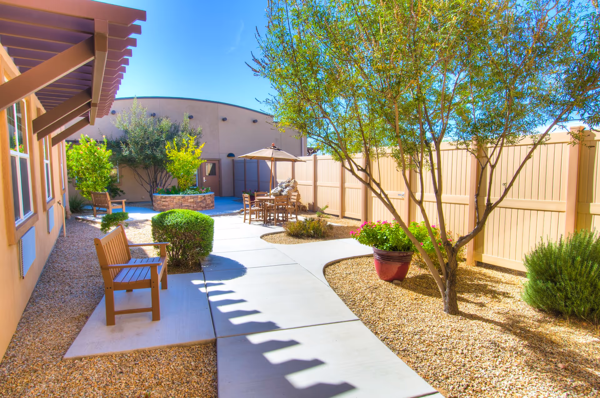 Outdoor courtyard area with a concrete walkway, wooden benches, a table with chairs and an umbrella, surrounded by trees, bushes, and a beige privacy fence under a clear blue sky.