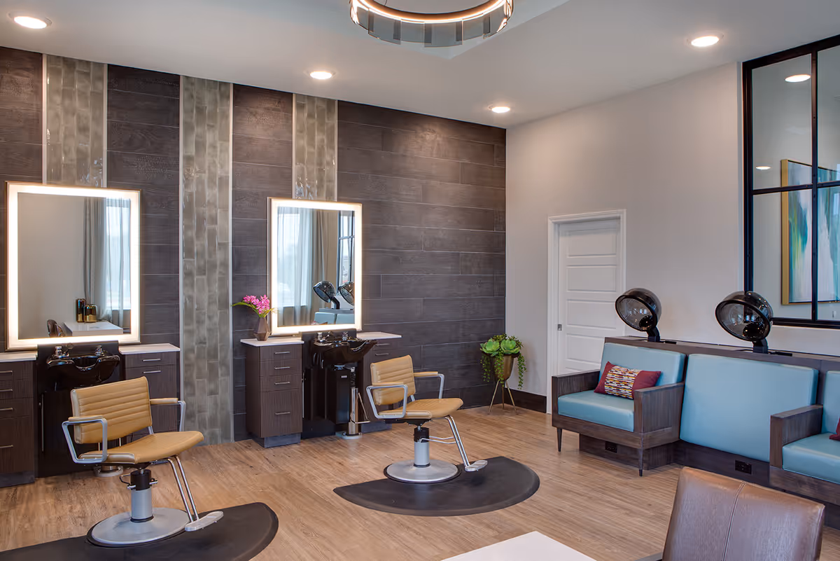 Interior view of a modern salon area in a senior living facility with two salon chairs in front of illuminated mirrors and wash basins, a seating area with two blue cushioned chairs and hair dryers, wooden flooring, and decorative plants.
