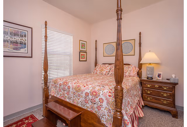 Cozy bedroom with a four-poster wooden bed dressed in floral bedding, a bedside table with lamp, framed wall art, and a window with blinds.