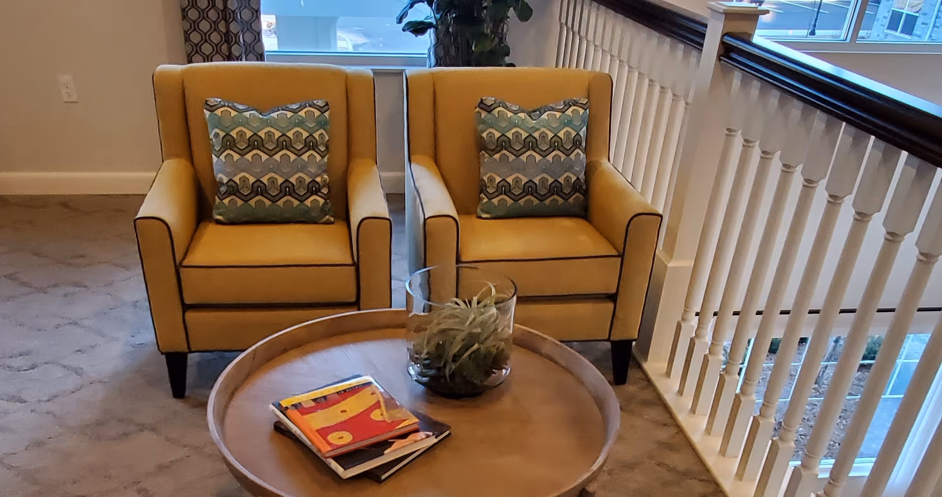Two yellow armchairs with patterned pillows beside a railing and a round coffee table holding magazines and a small plant in an interior seating area.