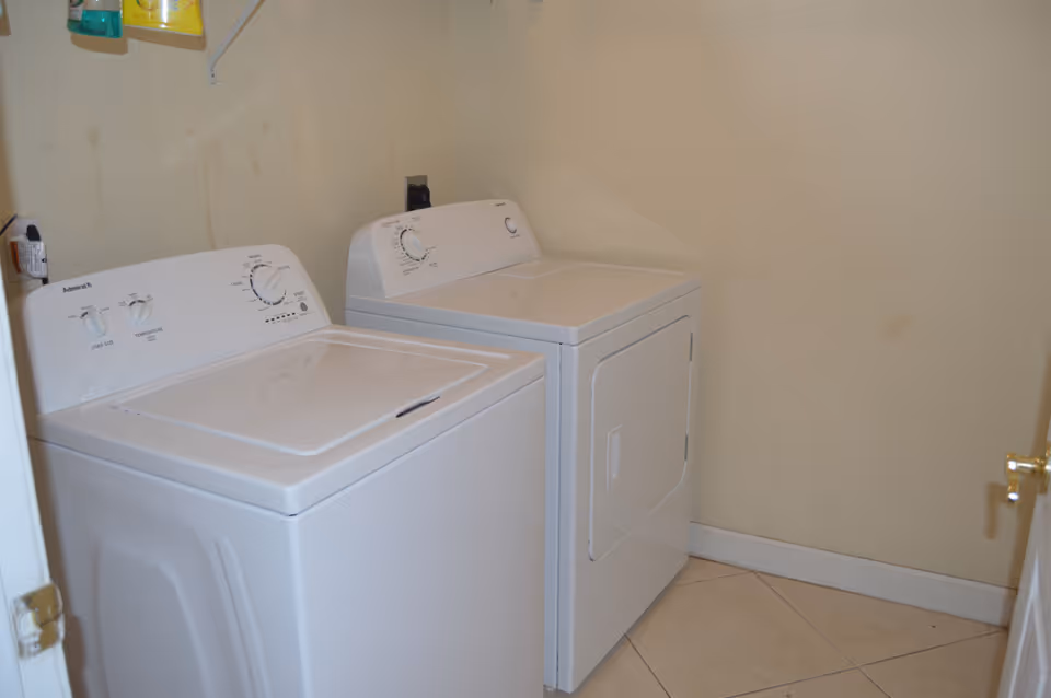 A small laundry room with a white top-loading washing machine and a white front-loading dryer placed side by side against a beige wall. There are some cleaning supplies on a shelf above the machines.