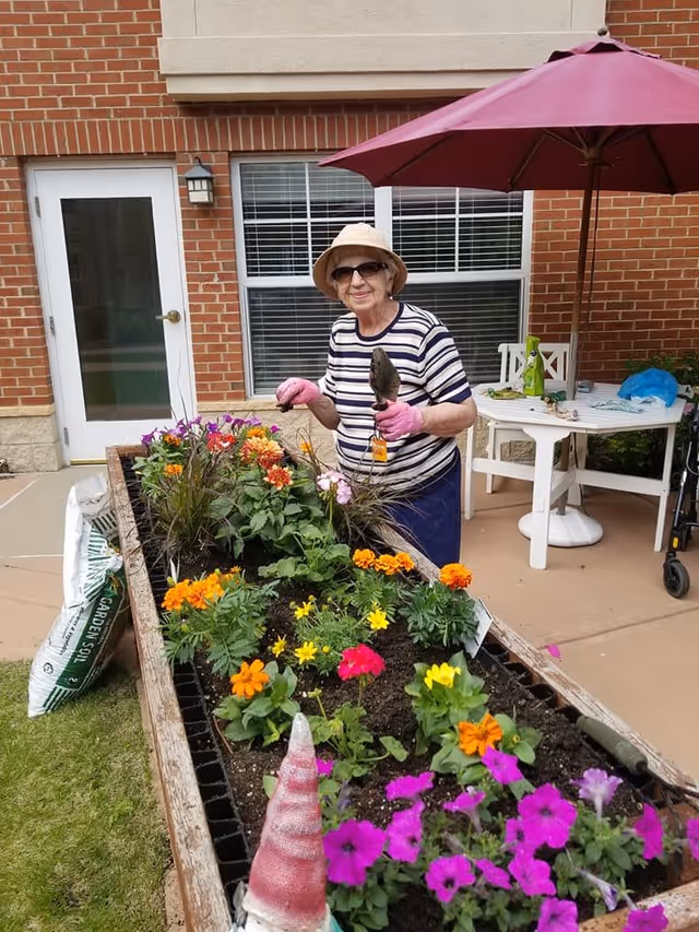 An elderly woman wearing a striped shirt, hat, sunglasses, and pink gardening gloves is tending to a raised garden bed filled with colorful flowers. She is holding a small gardening trowel and smiling. The scene is outdoors on a patio area with a brick building in the background, a white door, windows, a white table with a red umbrella, and gardening supplies nearby.