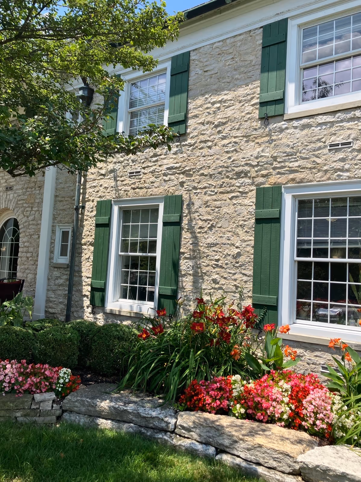 Stone exterior of a building with green window shutters, white-framed windows, and colorful flowerbeds in front.