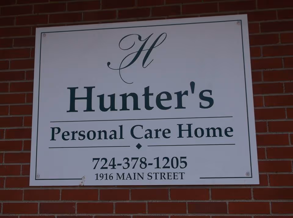 A white rectangular sign mounted on a red brick wall displaying the name and contact information of Hunter's Personal Care Home, including a phone number and address at 1916 Main Street.